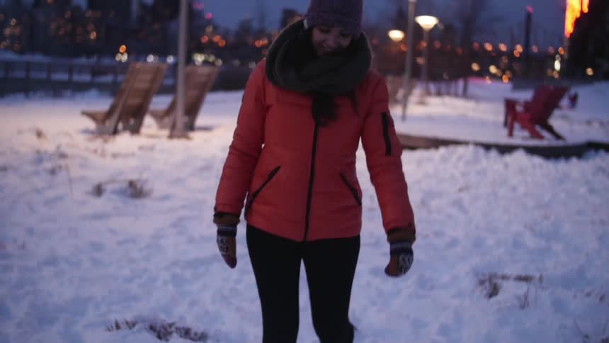 A woman in winter hat and coat kicks snow around at night