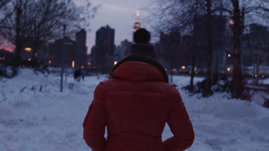 Woman in red jacket walks a snow covered path in the city at sunset
