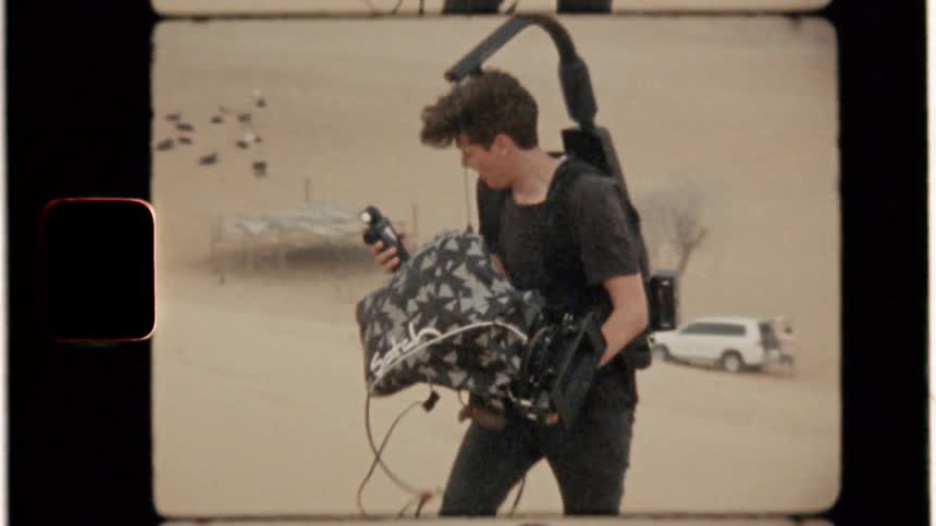 A cameraman wearing a microphone and harness walks through the sand