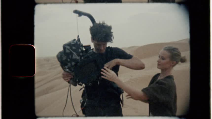 A woman helps a cameraman secure his harness and equipment in the desert
