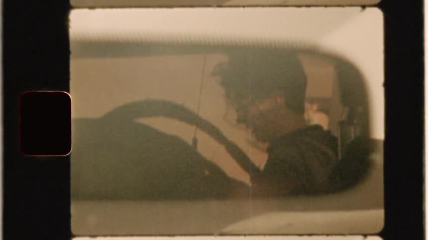 A car's rear view mirror shows a cameraman with a microphone on his pack