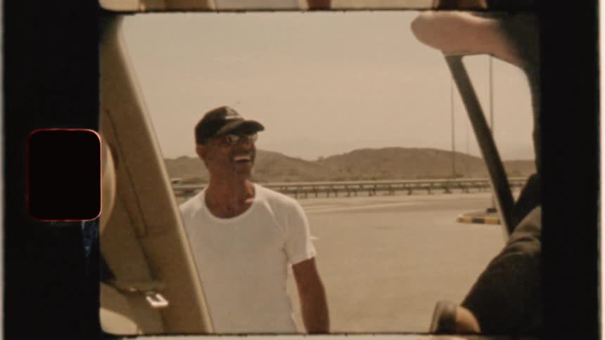 A man laughs at another who is balancing on a car door