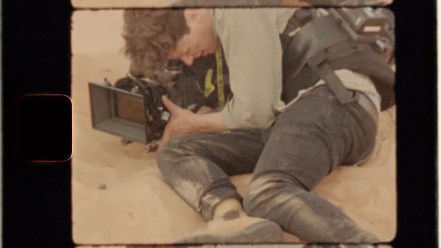 A cameraman sitting on a sand dune is covered in sand as he films during the day