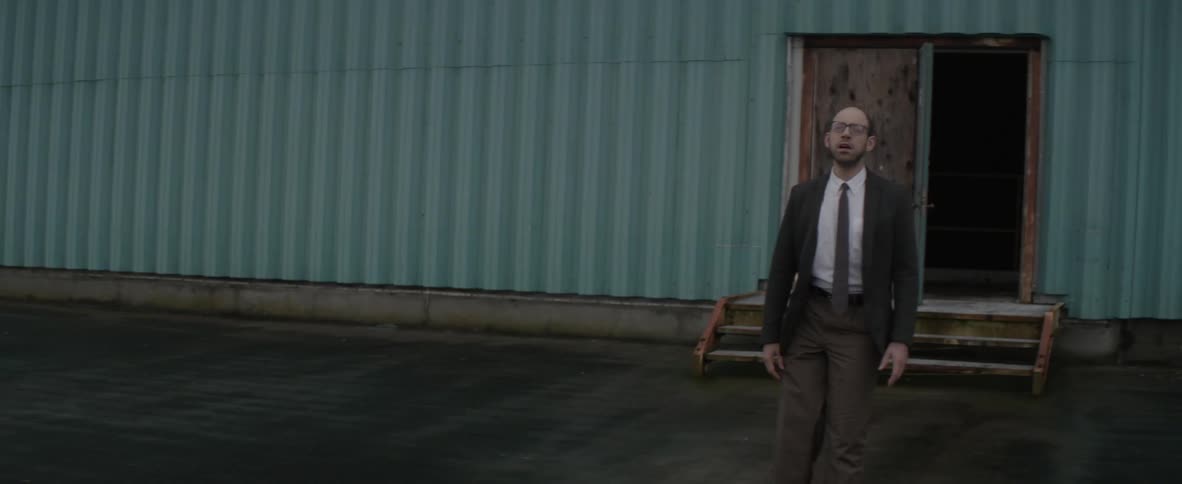 A man with a limp walks in a parking lot outside a warehouse and gazes in the distance