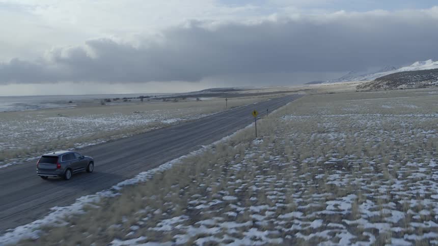 A car travels on a rural road passing fields dotted with snow