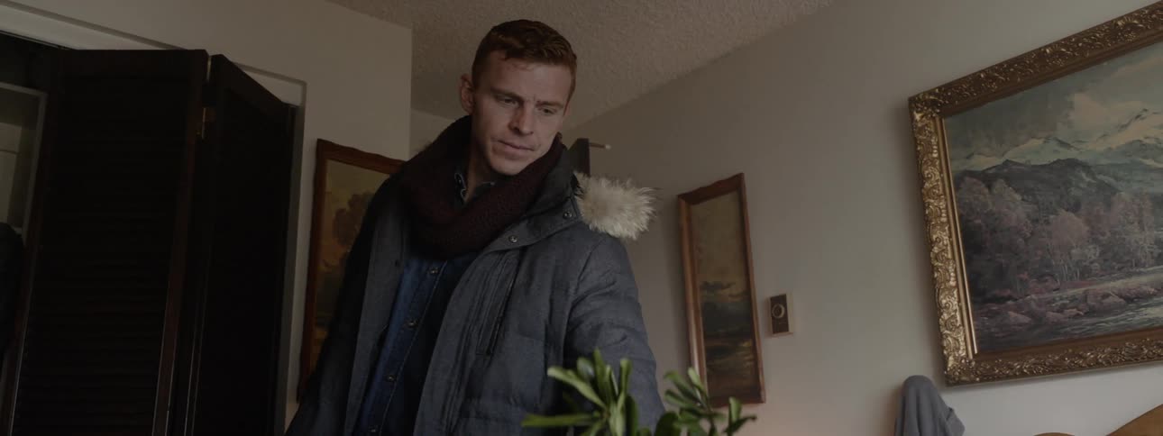 Man stands in a bedroom wearing a coat and examines a plant