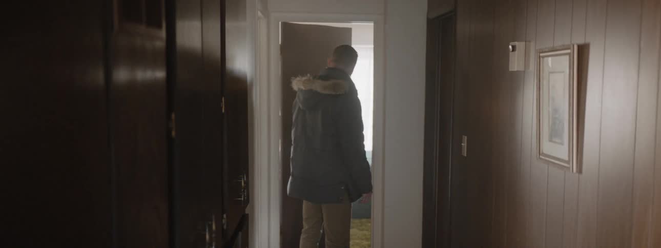 Man wearing a coat walks down a hallway and pushes open a door