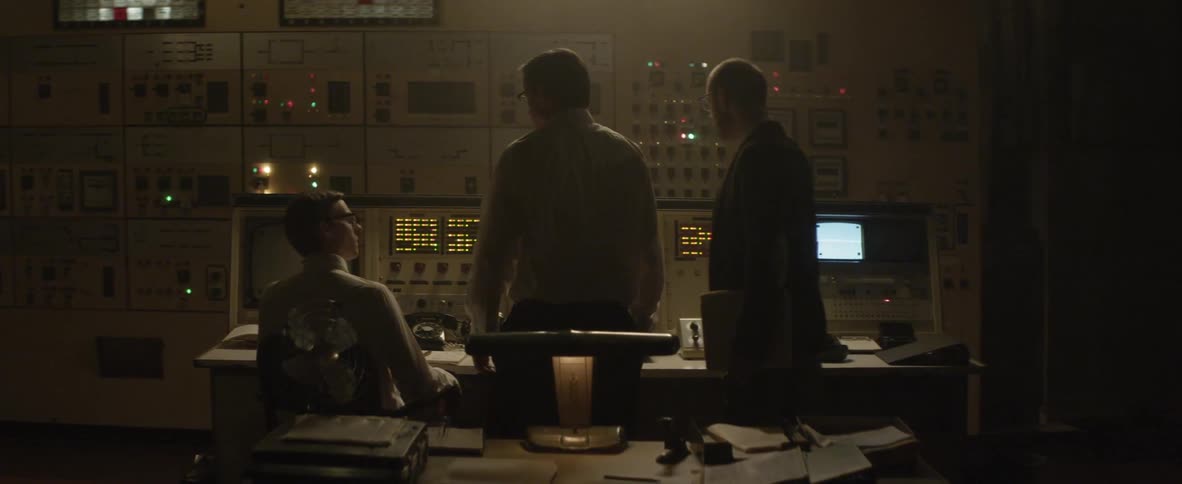 Several men talk to each other in front of a switchboard