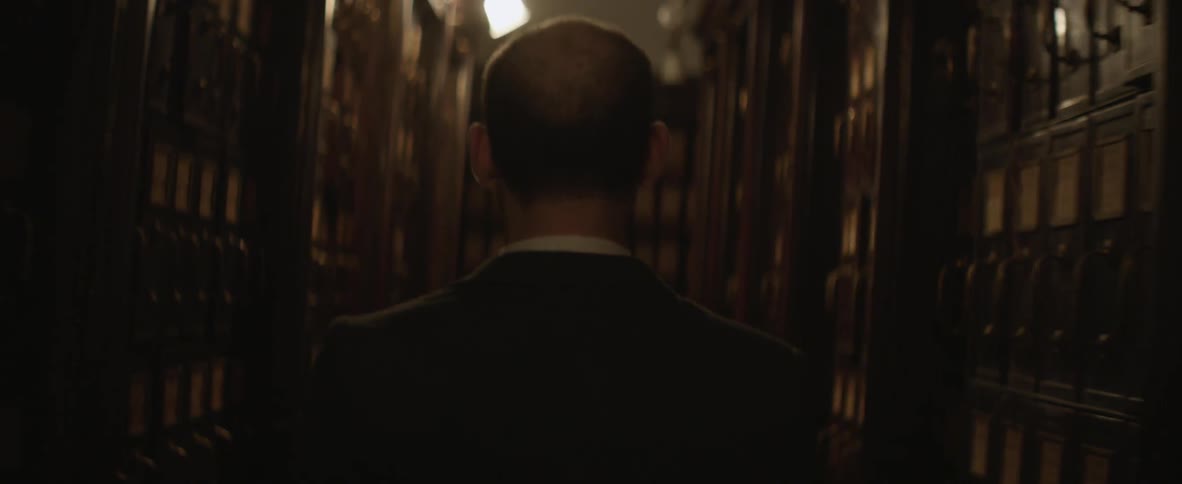 A man walks down a long aisle of tall bookshelves in a dimly lit library