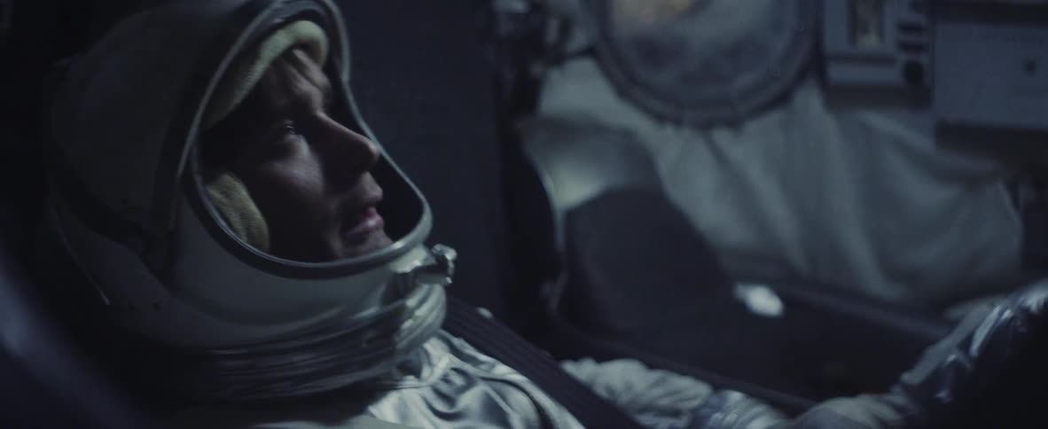 A stressed astronaut wipes his face while seated and wearing a spacesuit