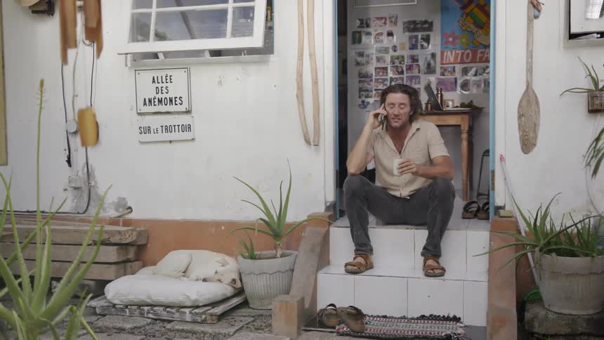 Man sits on a porch outside a small shop talking on a phone