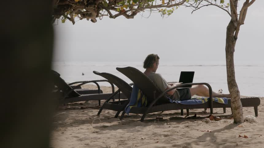 Man sits on a lounge chair at the beach typing on a laptop