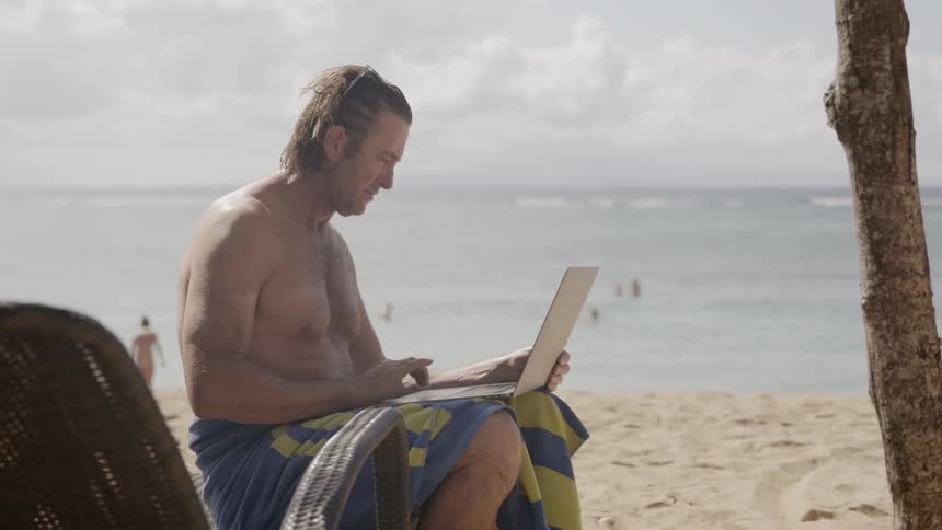A man sits on a beach chair working on his laptop