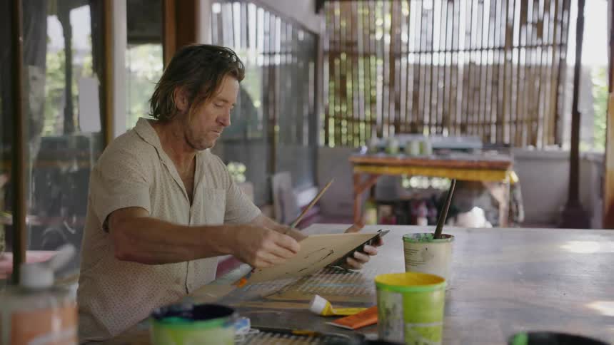 Man touches his cellphone as he paints on a small canvas
