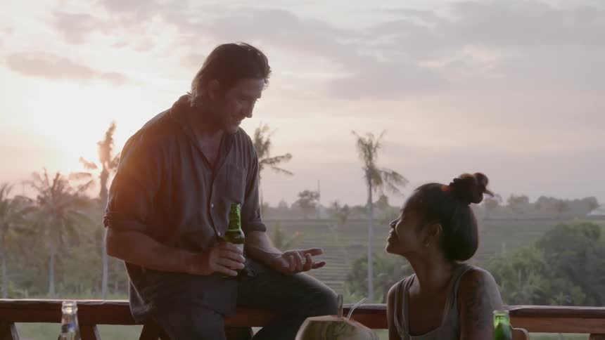 A man sits on a deck railing holding a beer and talking