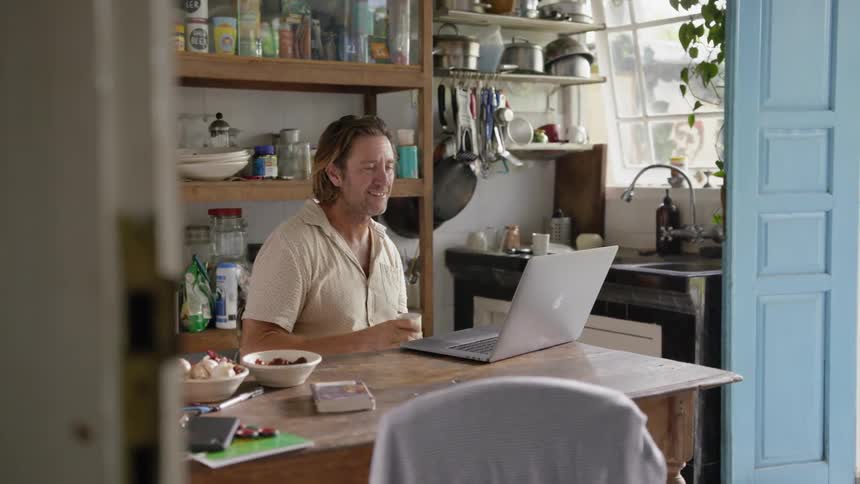 A man is ending a video call as he sits at a table in a kitchen in daylight