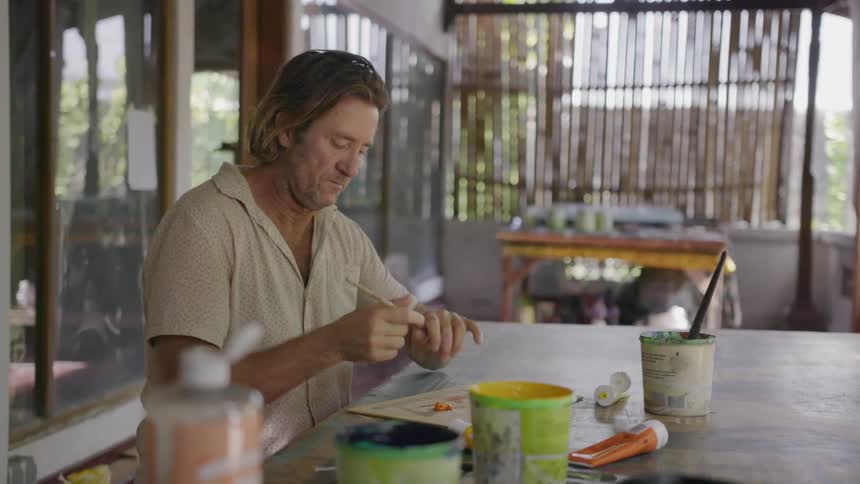 A man is mixing paint with a brush as he sits at a table outside in daylight