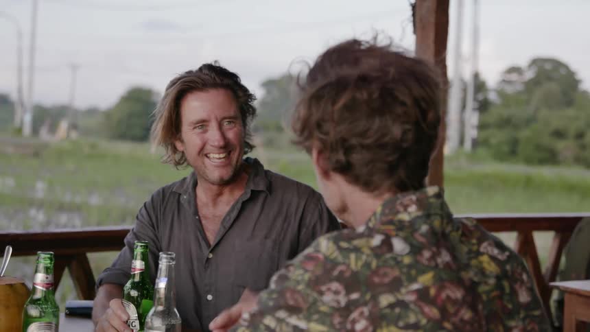 Two men sit at a table on a balcony smile and talk