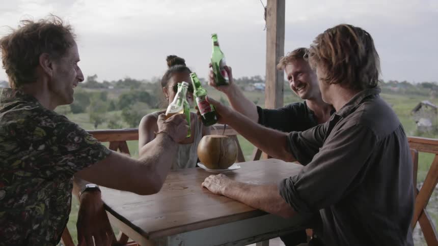 Four friends toast with beer bottles while sitting on a deck
