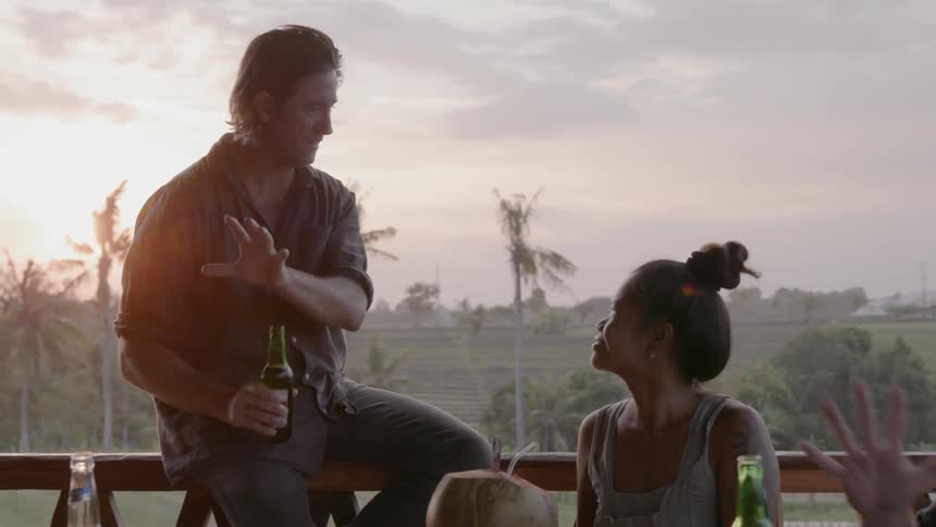 A man sits on a deck railing holding a beer bottle and talks with a woman