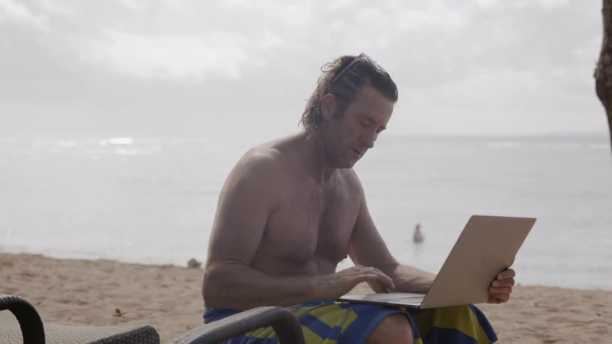 Man works on a laptop as he sits on the beach