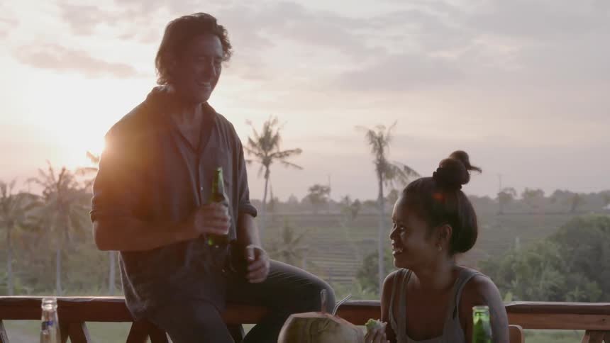 Man sits on a balcony railing talking to a woman who is sitting at a table