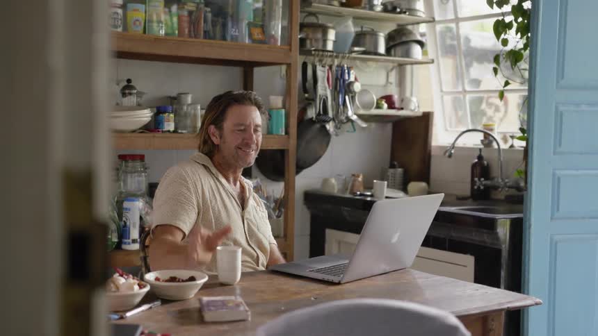 A man puts his mug down and laughs at something on a laptop