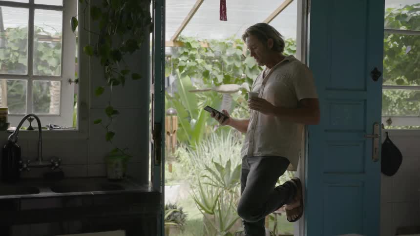 Man drinks a beverage and looks at his phone while leaning in a doorway