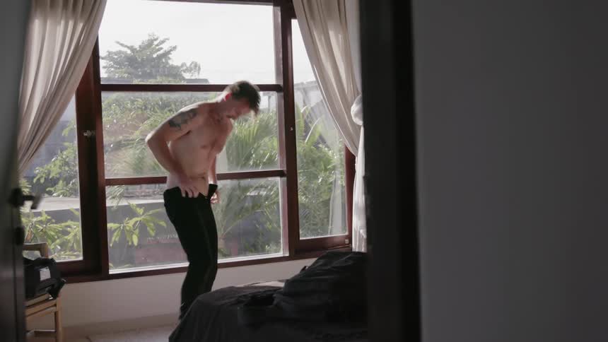 Man pulls up his pants as he stands shirtless in front of a window
