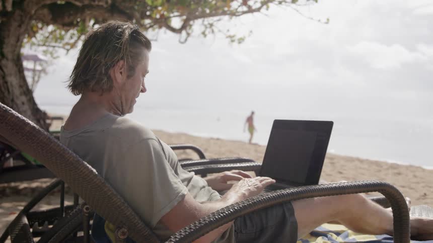 Man works on a laptop while sitting in a lounge chair at the beach