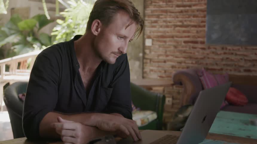A man is looking at his laptop and smiling as he sits in a patio in daylight