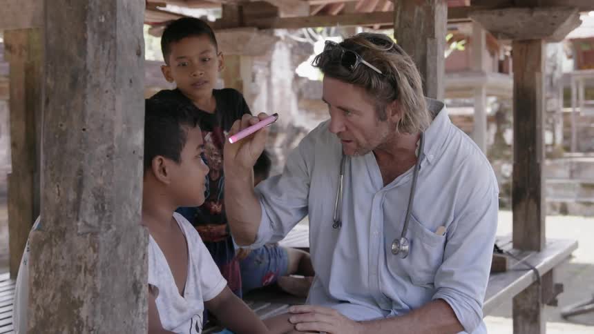 A doctor examines a young boy's eyes as another boy watches and laughs