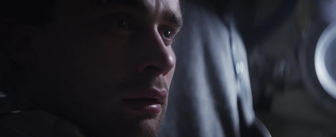 A close shot of a man in a space suit as he removes his helmet and sits pensively