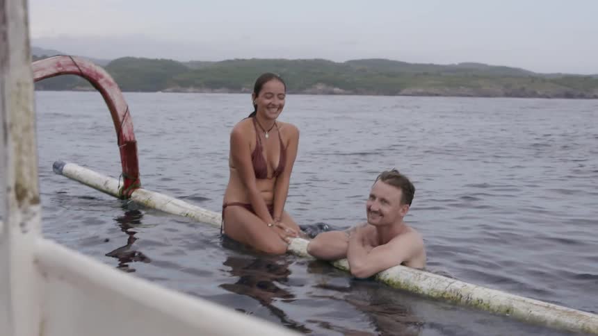 Man holds onto a metal log in the water as a woman sits on it