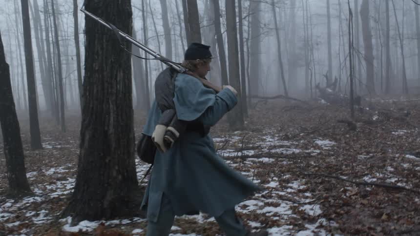 Man wearing a long coat and carrying a rifle walks in the forest