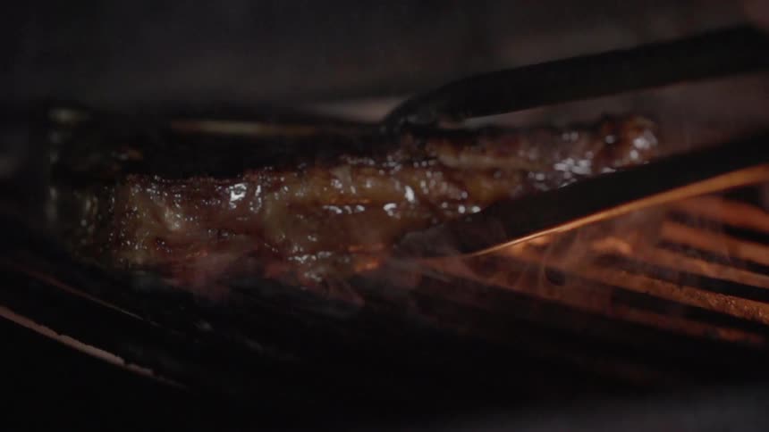 A steak on a flaming grill is picked up by tongs