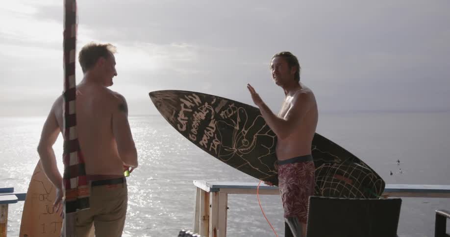 Man holds a surfboard as he talks to another man near the ocean