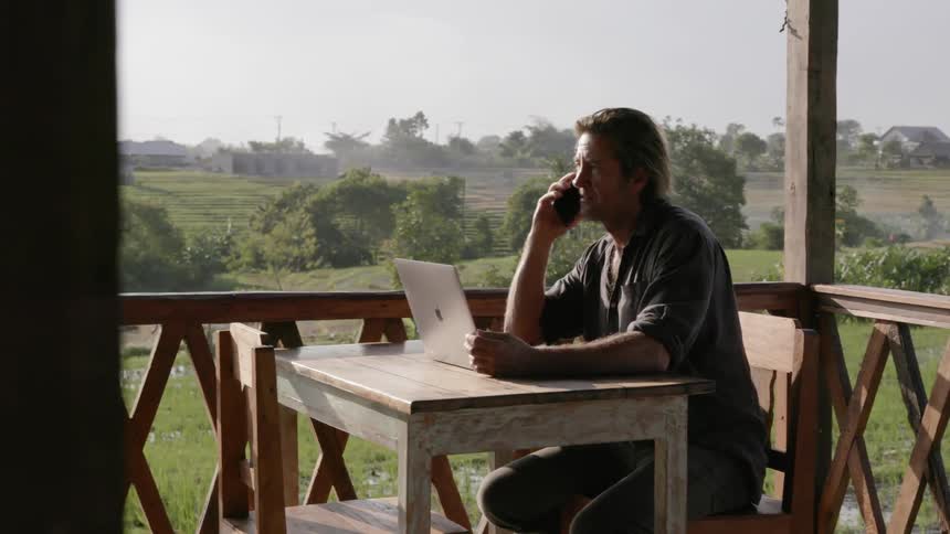 Man sits at a table on a balcony talking on a phone