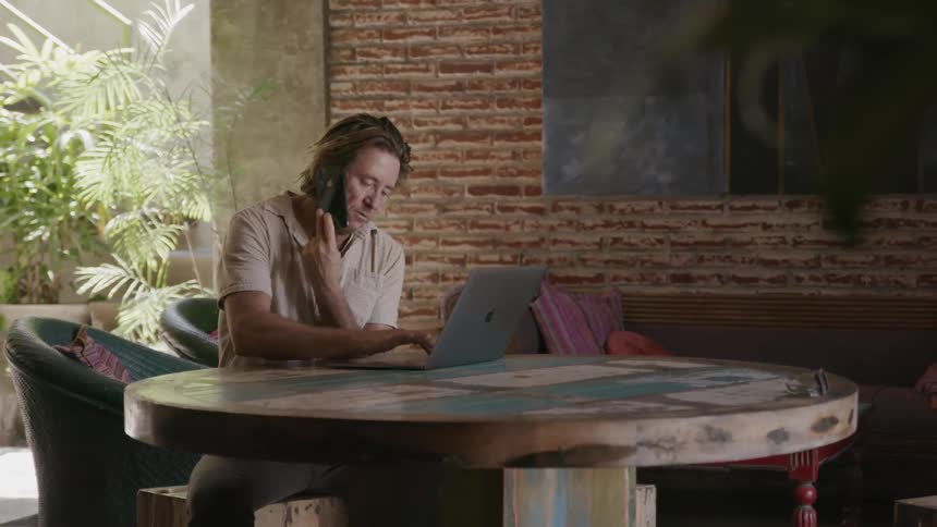 Man sits at a table talking on a phone and looking at a laptop