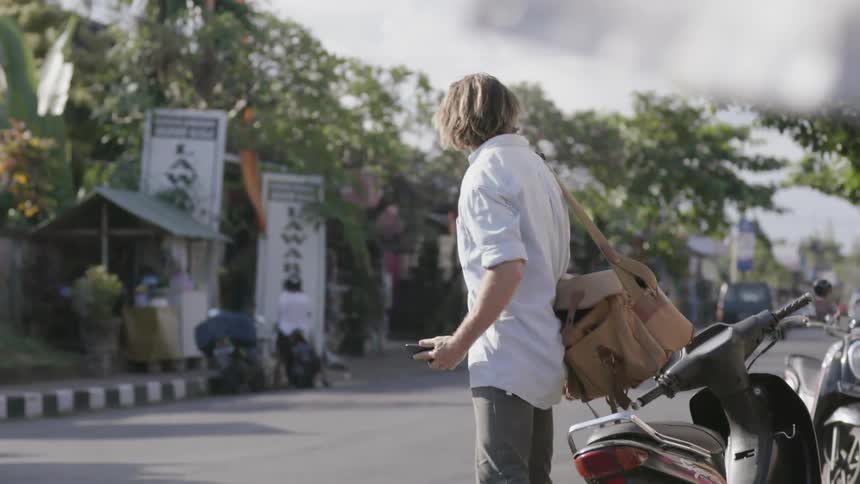 A man is picking up his pack and crossing a street outside in daylight