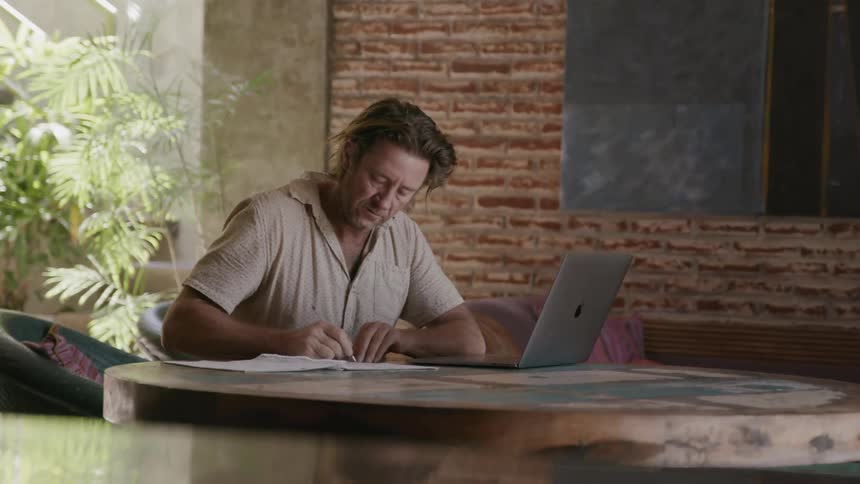 Man types on a laptop and writes in a notebook in a cafe