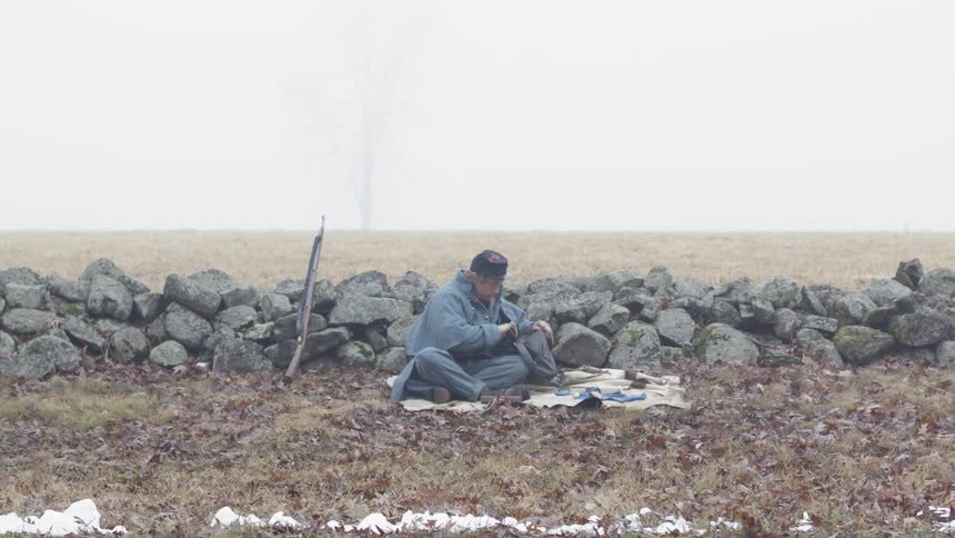 A union soldier sits on a blanket near a stone wall and packs his supplies