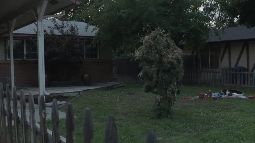 A person wearing leaves as camouflage walks towards the home