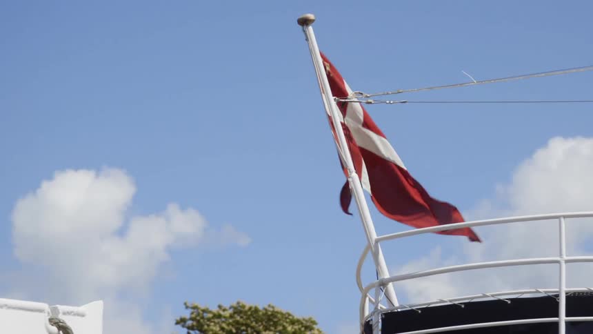 The flag of denmark is waving on a pole at the bow of a ship on a sunny day