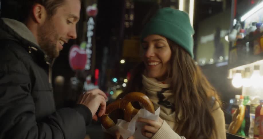 A woman takes a pretzel from a city food cart and turns to share with a man