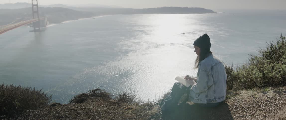 A woman sits on a ridge overlooking the ocean and writes in a journal