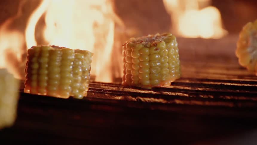 Corn on the cob is turned over on a flaming grill using tongs