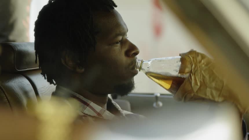 Man sits in a vehicle and drinks from a large bottle in a brown bag