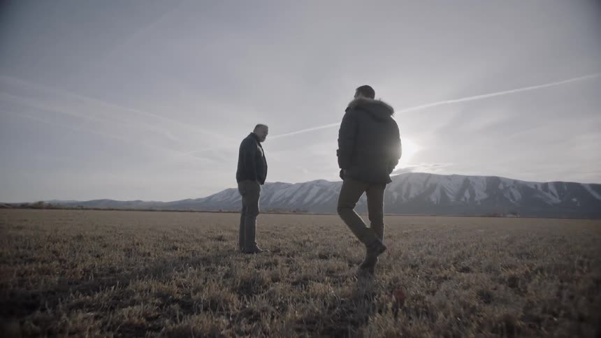 Two men walk in a rural field near mountains