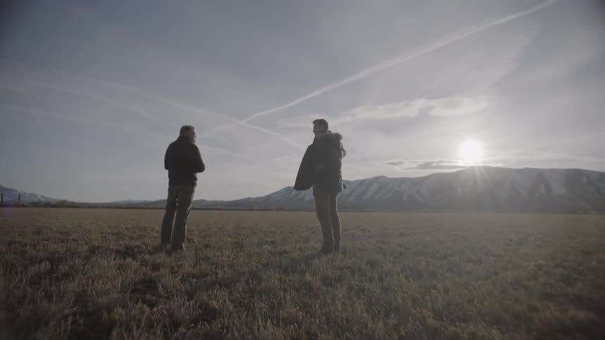 Two men stand in a rural field near mountains
